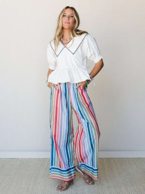 A bright, expressive summer outfit pairing a white peplum blouse with an oversized sailor collar against wide-leg multicolor stripe pants — the kind of look that feels equally at home on a boardwalk or at brunch.