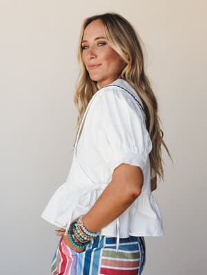 A model shot over the shoulder in a white peplum blouse with puff sleeves and tie waist, paired with bold multicolor striped pants and a stack of beaded bracelets — the kind of outfit that looks effortless but is clearly very much on purpose.