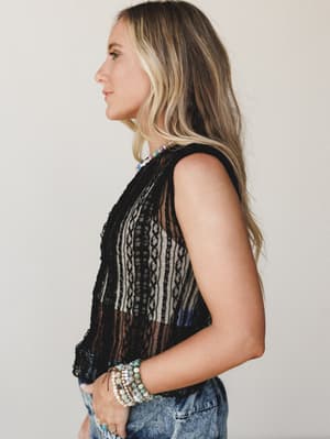 A side-profile shot of a model in a sheer black lace sleeveless top paired with acid wash jeans, styled with stacked beaded bracelets and a colorful beaded necklace — effortlessly boho with an edge.