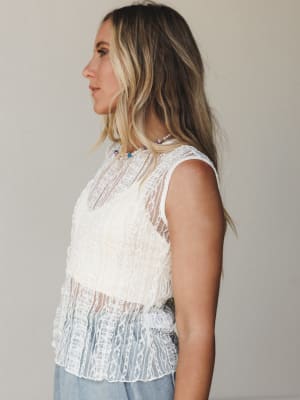 A sleeveless off-white lace top with sheer paneling and a scalloped hem, styled with light wash denim and a delicate beaded necklace — shot in side profile against a soft neutral backdrop with an easy, romantic feel.