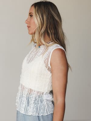 A sleeveless off-white lace top with sheer paneling and a scalloped hem, styled with light wash denim and a delicate beaded necklace — shot in side profile against a soft neutral backdrop with an easy, romantic feel.