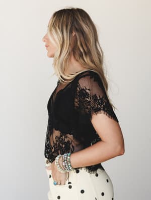 A side-profile shot of a model in a black sheer lace crop top with scallop-edge sleeves, layered over cream polka dot bottoms, styled with stacked stone and pearl bracelets — romantic and a little undone in the best way.