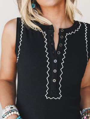A close-up detail shot of a black ribbed sleeveless top with white rickrack trim framing a button-front placket, styled with stacked beaded bracelets and turquoise drop earrings for a collected, boho everyday look.