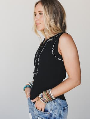 A side-profile shot of a model in a black sleeveless henley tank with white zigzag stitching, paired with light wash jeans and a stack of colorful beaded bracelets — casual and personal, like she just threw it on and it happened to be perfect.