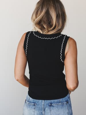 Back view of a black sleeveless knit tank with white zigzag contrast stitching along the neckline and shoulder seams, paired with light wash jeans against a soft grey studio backdrop.