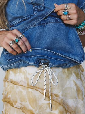A close-up detail shot capturing the hem of a denim vest layered over a tie-dye skirt, with both hands adorned in stacked silver and turquoise rings — the kind of styling that looks effortless but clearly isn't accidental.