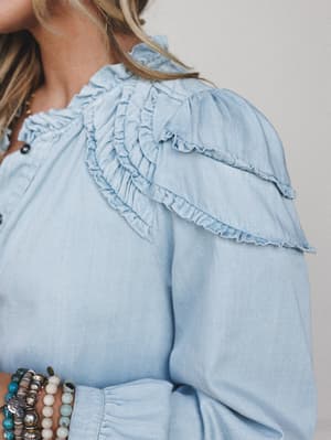A close-up detail shot of a light wash denim top with layered ruffle trim across the shoulder and yoke, styled with a stack of mixed beaded bracelets — romantic and a little western at the same time.