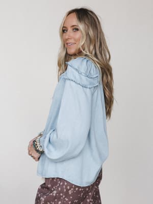 A light denim ruffle-trimmed blouse photographed from a three-quarter back angle, styled with a floral print bottom and stacked beaded bracelets — relaxed and a little romantic, the kind of top that makes a casual day feel intentional.