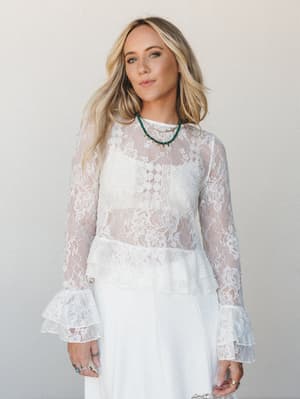 A sheer white floral lace long-sleeve top with tiered ruffle cuffs layered over a white dress, styled with a green beaded necklace and stacked rings — romantic and intentional, like something you'd wear to say 'I dressed for this moment.'