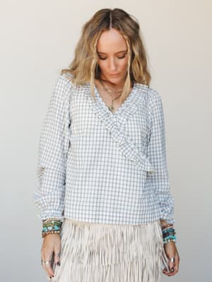 A ruffle-trimmed wrap blouse in a soft blue-grey gingham sits effortlessly on a model styled with stacked turquoise bracelets and a fringe skirt — the kind of outfit that feels put-together without trying too hard.
