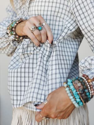 A close-up detail shot capturing the knotted hem of a black-and-white plaid ruffle blouse styled over a cream fringe skirt, layered with stacked turquoise, beaded, and silver bracelets and statement rings — the kind of accessory moment that makes the