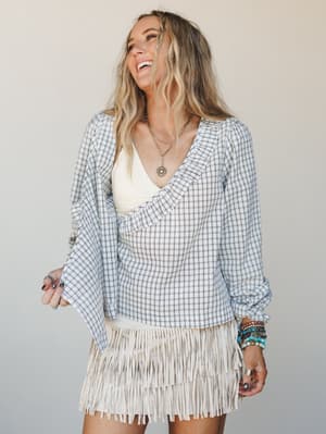 A laughing model layers a light blue plaid ruffle-trim blouse over a cream knit bralette, paired with a cream fringe mini skirt and stacked turquoise bracelets — the whole look has that effortless, free-spirited energy of someone who dressed for hers
