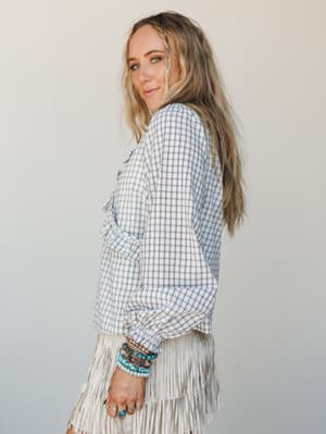 A relaxed side-profile shot of a woman in a cream and black plaid ruffle blouse paired with a cream fringe skirt, layered with turquoise and stone bead bracelets — the kind of outfit that feels effortless but clearly thought through.
