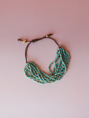 A multi-strand turquoise and gold seed bead bracelet with an adjustable brown cord closure, laid loosely on a dusty pink surface — the kind of piece that looks like it came home with you from a market you can't stop thinking about.