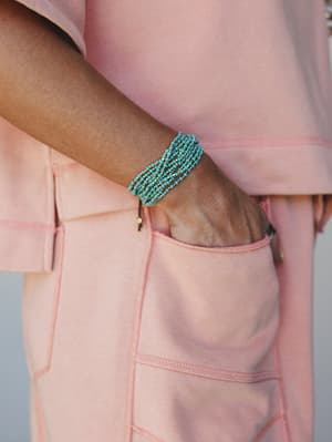 A close-up of a wide turquoise and gold beaded cuff bracelet worn on the wrist, hand tucked into the pocket of a soft pink cargo-style bottom — the color contrast does all the talking.