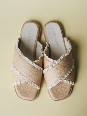 A pair of woven raffia slide sandals with frayed lace trim along the crossover straps, shot flat lay against a soft sage-green background — the kind of sandal that makes you want to book a trip somewhere warm.