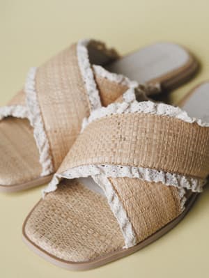 A pair of woven raffia slide sandals with crisscross straps and delicate crochet lace trim, resting casually on a soft sage-yellow surface — the kind of shoe that makes a sundress feel complete.