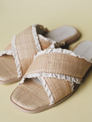 A pair of woven raffia slide sandals with crochet-trimmed crossover straps, shot close-up against a soft yellow-green backdrop — the kind of sandal that makes every summer outfit feel a little more intentional.