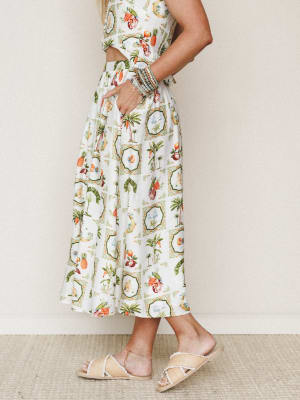 A cropped side-profile shot of a model in a breezy tropical-print co-ord set — a midriff-baring crop top and full midi skirt — paired with natural raffia slide sandals and a stack of beaded bracelets, shot against a soft neutral studio wall.