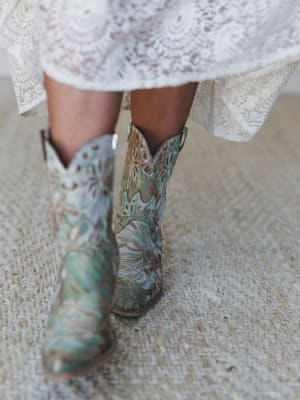 A close-up shot of intricate embroidered cowboy boots in sage and turquoise paired with a flowy lace dress hem — the kind of combination that feels equally at home at a barn wedding or a weekend festival.