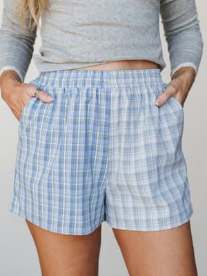 A relaxed mid-shot of plaid pull-on shorts in soft periwinkle blue paired with a grey long-sleeve top, hands tucked in pockets — the kind of outfit that blurs the line between lazy morning and actually leaving the house.