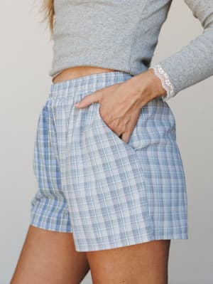 A close mid-shot of plaid pull-on shorts in soft blue and white paired with a grey cropped long-sleeve top with lace cuff detail — the kind of effortless at-home outfit that makes staying in feel intentional.