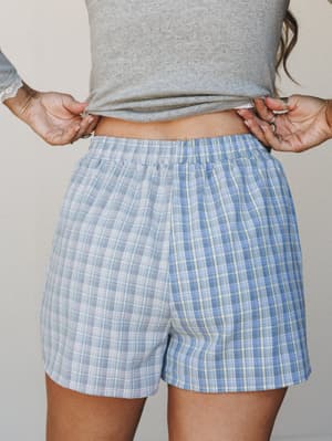 A close-up mid-shot of relaxed plaid shorts in soft blue and white with faint yellow grid lines, styled with a grey textured crop top — the kind of effortless combo that makes a lazy morning feel intentional.
