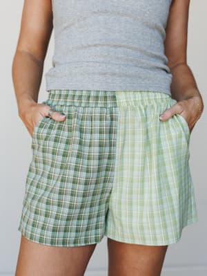A close-up mid-shot of relaxed plaid shorts in a clever two-tone colorblock — forest green on one side, soft lime on the other — worn hands-in-pockets over a gray pointelle tank, giving off easy weekend energy.