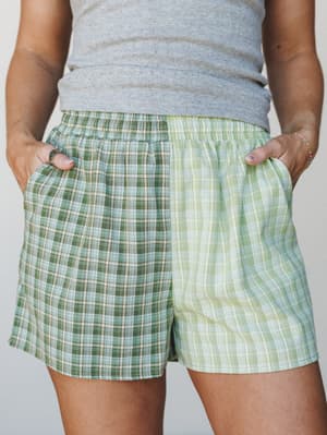 A close-up of color-blocked plaid shorts in two shades of green — one panel deep forest, one soft lime — styled with a grey pointelle tank, hands tucked in pockets, relaxed and easy.