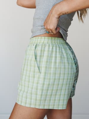 A close side shot of breezy plaid mini shorts in a soft sage and lime green colorway, styled with a cropped gray tee and boho jewelry — the kind of easy warm-weather outfit that looks effortless without trying.