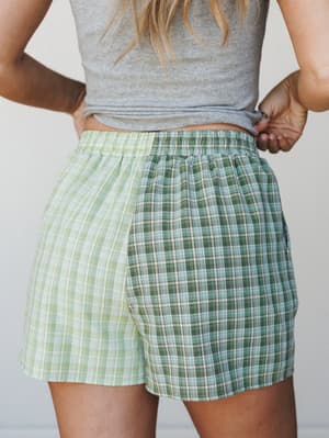 A close-up back view of color-blocked plaid shorts in two shades of green — light lime and deeper forest — paired with a casual gray crop top, shot against a clean neutral background.