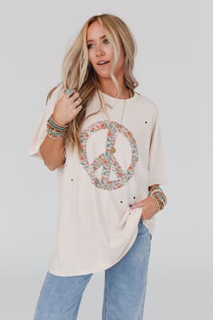 A woman with long blonde hair wears the Jude Oversized Peace Sign Graphic Tee in oatmeal, blue jeans, and colorful bracelets, standing against a plain white background with one hand in her pocket.