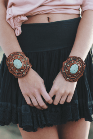 A person in a black skirt and pink top showcases two Aegean Boho Leather Cuff Bracelets in brown, featuring turquoise stones and metal accents—crafted from quality vegetable tanned leather for a perfect boho look.
