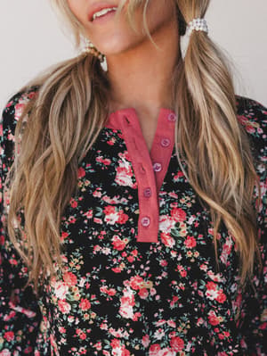 A close-up detail shot of a black floral top with a dusty rose henley placket and button detail — the kind of cozy, pretty thing you reach for without thinking twice.