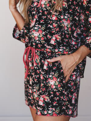 A close-up torso shot of a black floral PJ set with a coral drawstring waist, hand tucked into a side pocket — the kind of set that makes staying in feel like a whole mood.