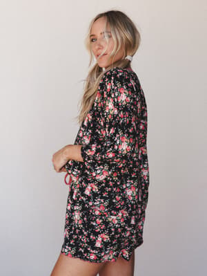 A floral print PJ set in black with rosy blooms — shot in three-quarter profile against a soft neutral wall, with that effortless 'morning but make it pretty' energy.