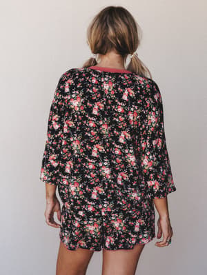 A matching floral top and shorts set photographed from the back — black base with pink and cream roses, worn with pearl hair clips and a relaxed, lived-in ease that blurs the line between pajamas and a really good Saturday outfit.