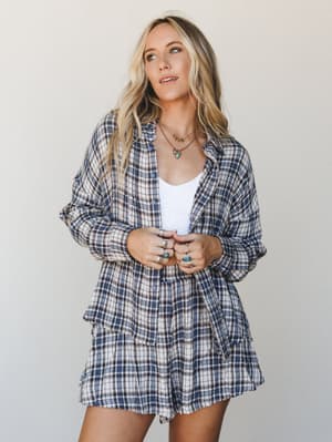A plaid matching set — oversized button-down shirt and relaxed shorts in navy and cream — worn open over a white tank with layered gold and turquoise jewelry, giving off that effortless 'rolled out of bed and still look good' energy.