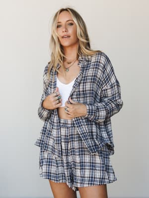 A matching plaid flannel set — oversized button-down shirt worn open over a white bralette with coordinating shorts — styled with layered gold and turquoise jewelry for an effortlessly lived-in look.