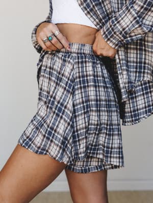 A close-up mid-shot of a matching plaid flannel short set — relaxed shorts paired with an open plaid shirt over a white crop top, styled with silver and turquoise rings for that effortless, lived-in feel.