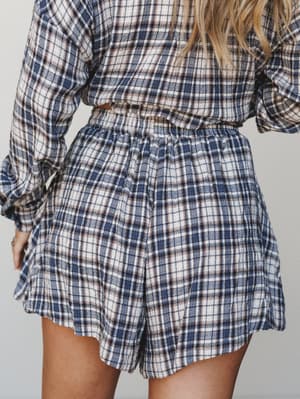 A close-up back view of a matching plaid set — flowy shorts and a long-sleeve top in a navy, cream, and rust plaid — soft, lived-in, and the kind of cozy you actually want to be seen in.