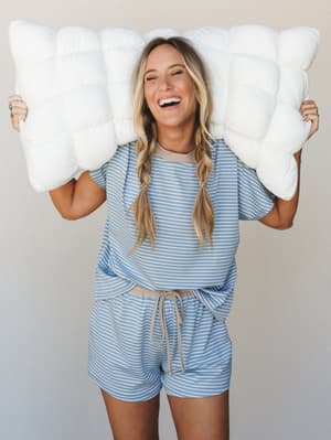 A laughing woman holds a fluffy white pillow above her head while wearing a matching striped short-sleeve top and shorts set in soft blue and cream — the kind of outfit that makes staying in feel like a whole personality.