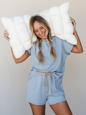 A smiling model holds a oversized puffy pillow above her shoulders while wearing a matching striped short set in soft denim blue and white with taupe trim details — the whole vibe is cozy morning energy, the kind where you're happy and slightly unhin