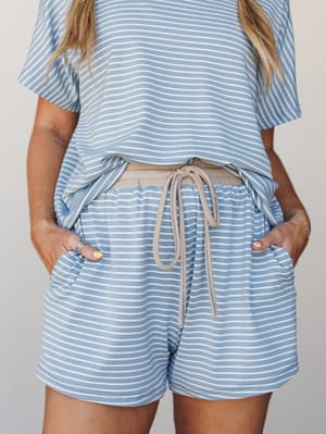 A close-up of a matching striped set — cropped tee and pull-on shorts in soft periwinkle blue and white stripes with a taupe drawstring waist, hands tucked in pockets like she's exactly where she wants to be.