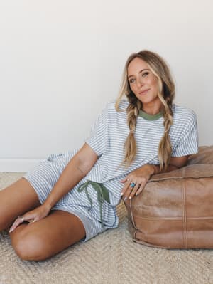 A relaxed, lived-in moment — model seated on a jute rug leaning against a leather pouf, wearing a matching striped tee-and-shorts set with soft olive contrast details and braided pigtails that feel effortlessly at-home.
