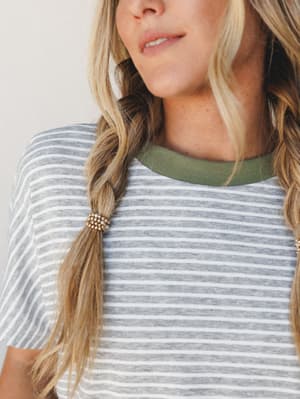 A close-up shot of a striped tee with a sage green ribbed crew neck, worn by a blonde model with loose braids tied with gold beaded hair ties — casual and easy with just enough personality.
