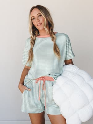 A relaxed matching stripe set in soft mint and white with cheerful coral accents — she's holding a fluffy white comforter like she just rolled out of the coziest morning, and somehow looks completely put together doing it.