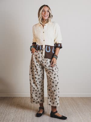 Woman wearing a cream shirt with brown horse shoe design and patterned pants against a plain background