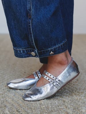 A close-up of silver metallic Mary Jane flats with double buckle straps and grommet detail, paired with dark wash wide-leg jeans — the kind of shoe that makes a quiet outfit feel like a whole moment.