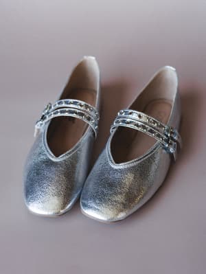 A pair of silver metallic Mary Jane flats shot from above against a muted mauve background — the double grommet straps give them just enough edge to feel special without trying too hard.
